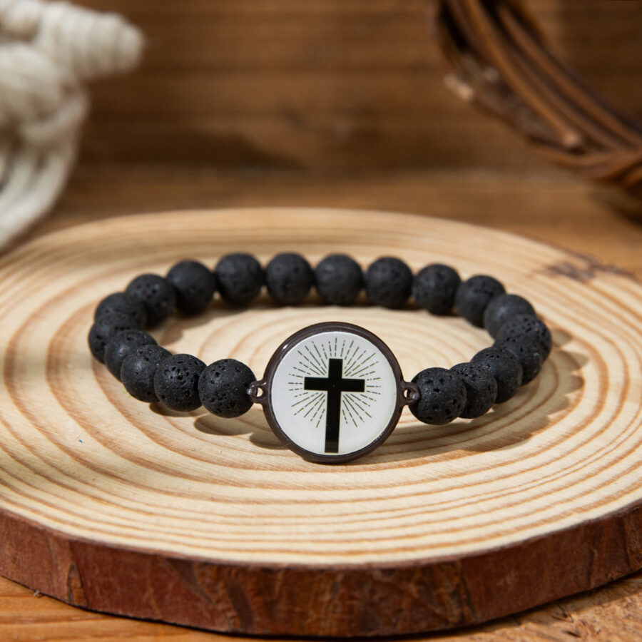 Black lava rock bracelet with a white, circular cross charm and sunburst design, set on a rustic wooden platter.