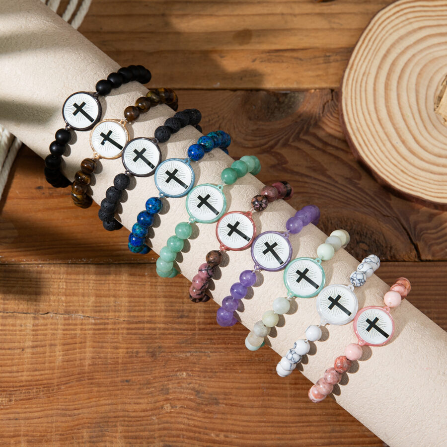 A collection of six different colorful natural stone bead bracelets with cross charms, arranged on a wooden surface. Colors include black, blue, pink, purple, and green.