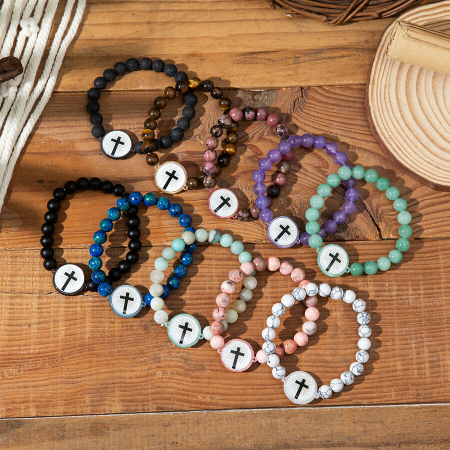 A collection of six different colorful natural stone bead bracelets with cross charms, arranged on a wooden surface. Colors include black, blue, pink, purple, and green.